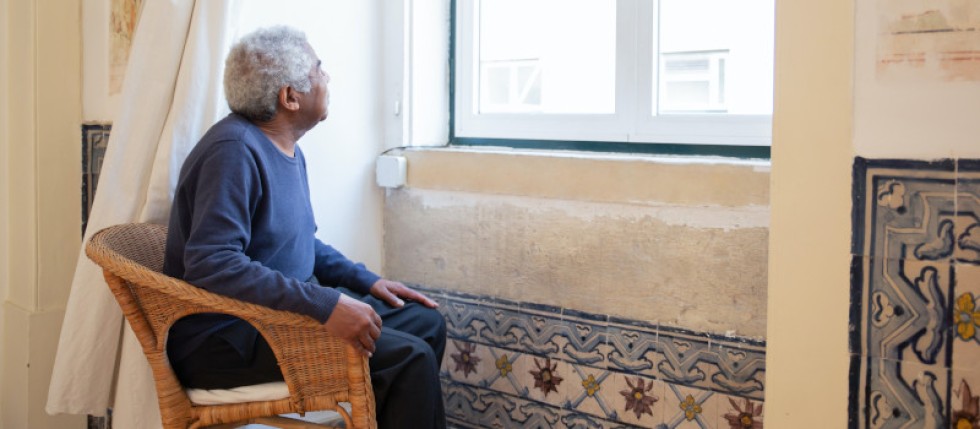 Senhor sentado, de cabelos brancos, à janela, espera apoio para obras em casa de idosos.