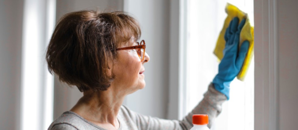 Mulher com óculos e luva azul limpa uma janela seguindo dicas para limpeza doméstica rápida.