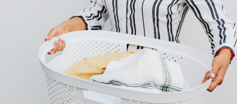 Mãos de mulher com camisa branca às riscas azuis a segurar cesto de roupa branco e a pensar em como tirar manchas amarelas da roupa guardada.