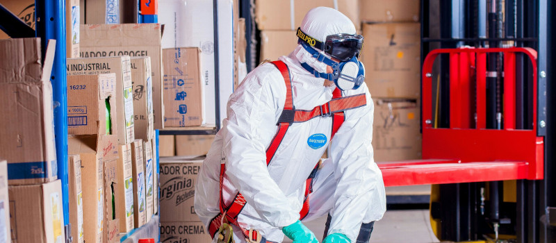 Trabalhador com farda branca, óculos de proteção e luvas cumpre regras de saúde e segurança no trabalho a mexer em produtos tóxicos, rodeado por caixas de papelão.