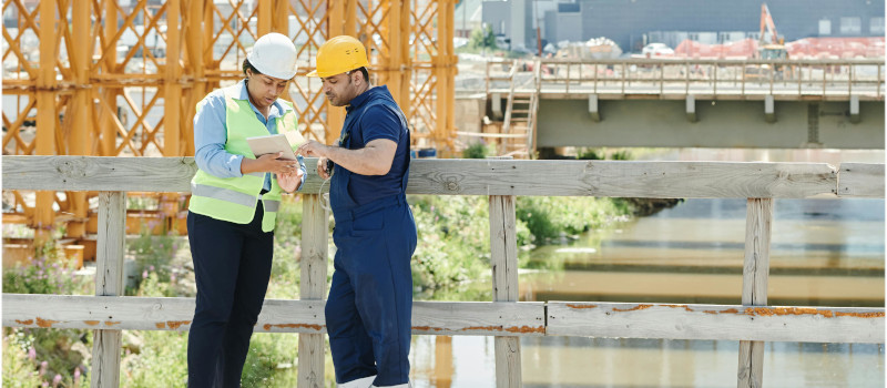 Mulher e homem com capacetes na cabeça em obra de construção analisam normas de segurança no trabalho.