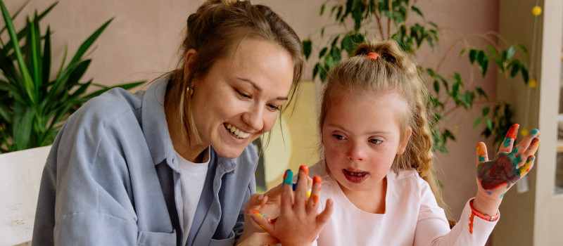 Menina loira com Síndrome de Down, tinta nos dedos e totós na cabeça, a receber apoio de terapeuta sorridente através de um subsídio de educação especial para terapia da fala.