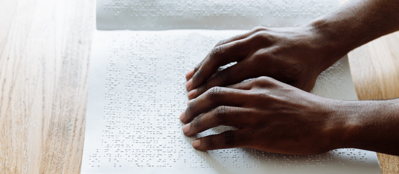 Mãos de jovem de pele negra a ler livro em braille em cima de mesa bege usufrui de subsídio de educação especial.