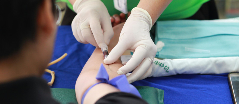 Mãos de enfermeiro com luvas brancas a espetar agulha no braço de homem de óculos para lhe tirar sangue.