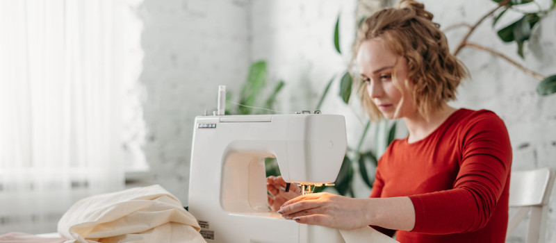 Mulher loura de camisola vermelha a coser à máquina vai pensando nos tipos de costura que existem.