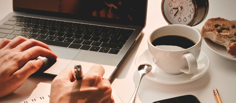 Mãos de homem jovem, uma sobre o computador, outra a escrever, com chávena de café ao lado e relógio em frente, à procura de ferramentas de gestão de tempo.