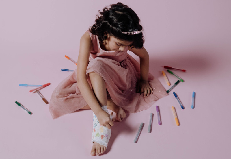 Menina morena com vestido cor-de-rosa em fundo também rosa com marcadores coloridos ao redor, vai pintando o gesso num pé enquanto a mãe pensa no seguro escolar.