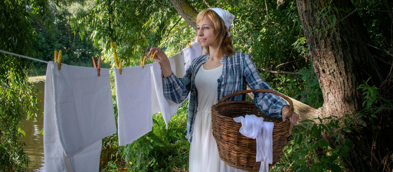Mulher sorridente de vestido branco e lenço branco na cabeça, com camisa aos quadrados azuis e brancos, segura cesto de vime enquanto apanha roupa branca de uma corda estendida ao ar livre.