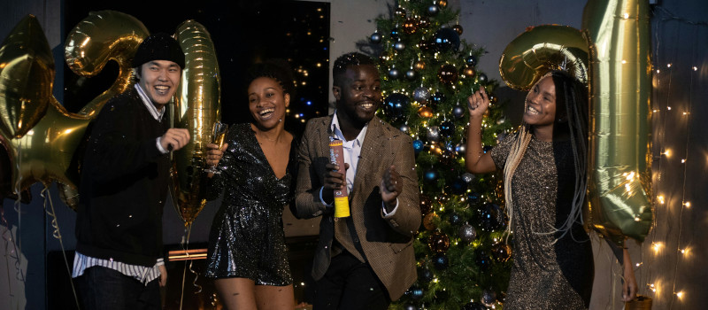 Festa de passagem de ano em casa com árvore de Natal e decorações douradas atrás e quatro pessoas sorridentes a celebrar: duas mulheres negras com roupas brilhantes, um homem asiático com gorro e camisola preta e um homem negro de blazer castanho.