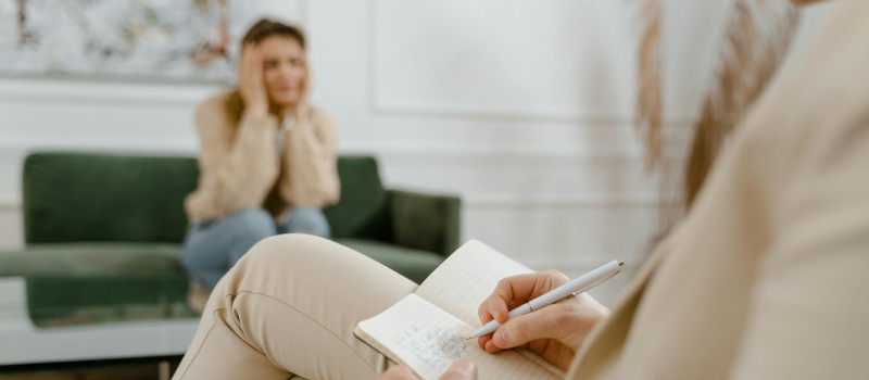 Mulher de fato bege a tomar notas enquanto estudante desfocada na imagem tem mãos na cabeça durante consulta do Cheque Psicólogo.