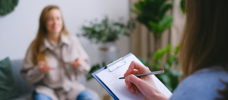 Mulher de camisa azul a tomar notas enquanto estudante desfocada fala com as mãos à frente do corpo durante consulta do Cheque Psicólogo.