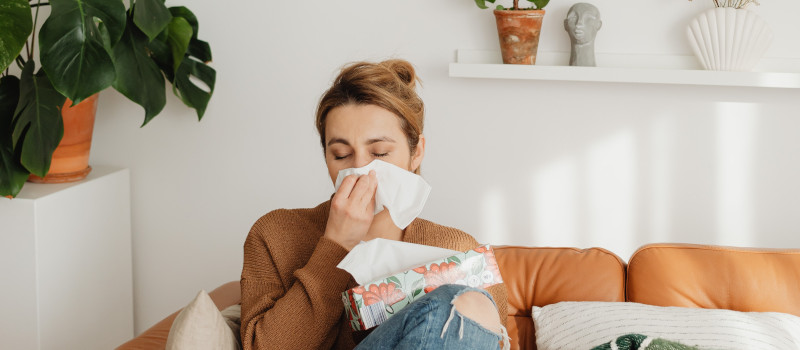 Mulher sentada num sofá castanho, com uma camisola castanha e jeans, a pensar nos objetos que causam alergia em casa.