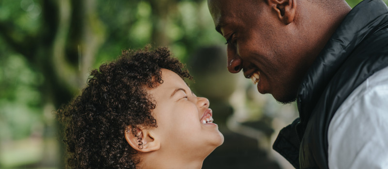 Homem de pele morena e criança de cabelo encaracolado sorriem a olhar um para o outro enquanto pensam porque mentem as crianças.