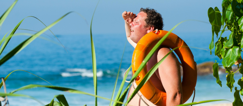 Homem na praia, com o rosto vermelho e em tronco nu, a olhar o horizonte com uma boia laranja ao ombro a pensar como tratar a alergia ao calor.