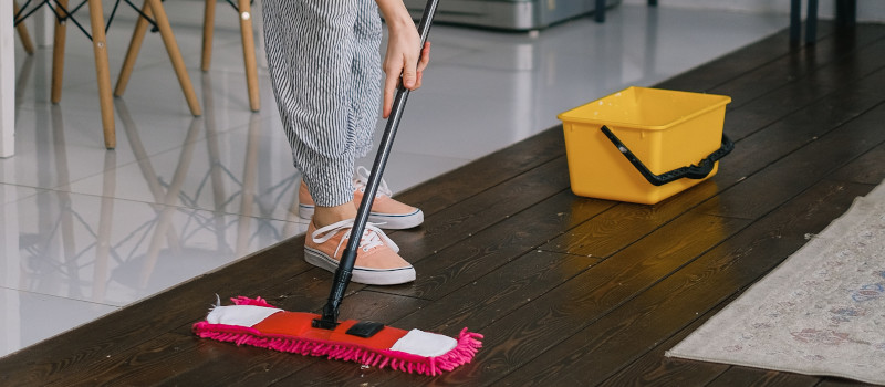 Mãos de mulher com calças às riscas e ténis cor de laranja seguram esfregona vermelha com um balde amarelo ao lado com um produto para limpar chão de madeira envernizado.