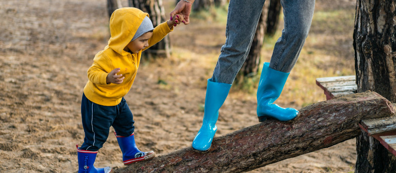 Pés de mulher com galochas azuis e calças de ganga em cima de um tronco a dar a mão ao filho bebé de camisola amarela e galochas azuis claras.