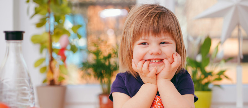 Menina sorridente, com as mãos a segurar o queixo e a mostrar os dentinhos, enquanto os pais pensam em saber como aumentar a autoestima das crianças