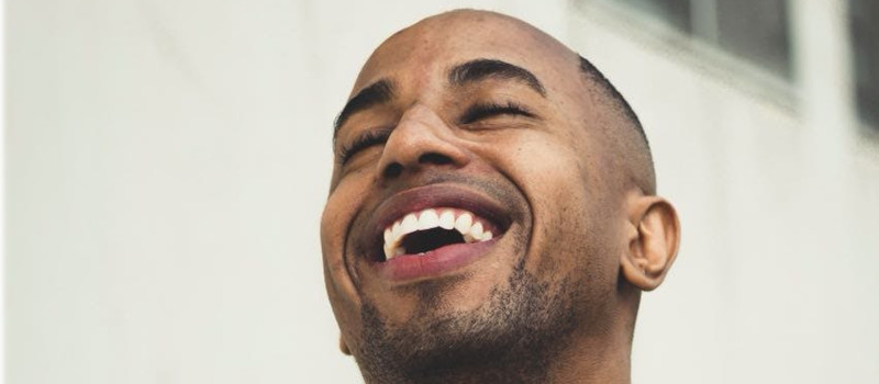 Homem moreno, com cabelo rapado e barba rasa, ri mostrando os dentes e como fazer terapia do riso.