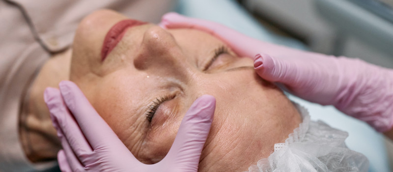Mulher idosa de olhos fechados e touca branca na cabeça, recebe terapia manual de mãos com luvas rosa em sessão de fisioterapia gerontológica.