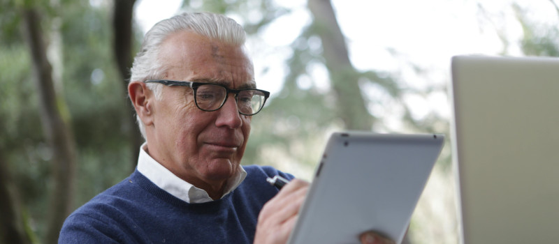 Homem idoso de cabelos brancos, óculos pretos, camisa branca e camisola azul a olhar para um tablet e a fazer a pesquisa 