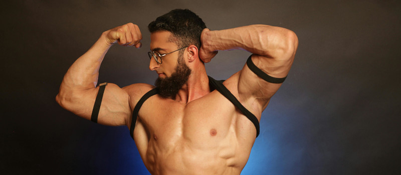 Homem musculado em tronco nu, de óculos e barba, a fazer pose com o braço e a olhar para os próprios músculos a ver resultados do bulking.