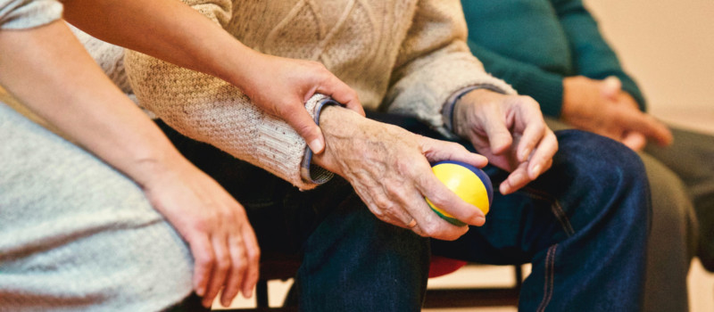 Mão de mulher com Estatuto do Cuidador Informal a segurar pulso de idoso que tem uma bola amarela na mão.