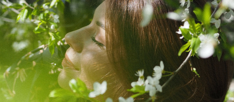 Mulher jovem de olhos fechados, cabelo castanho, respira o odor das flores num dos tipos de jardins sensoriais.