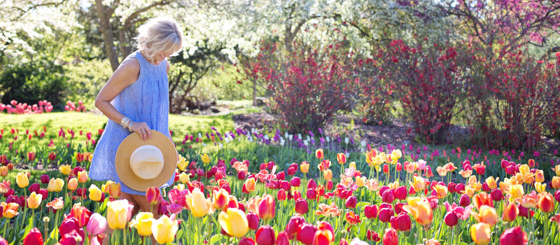 Mulher loura de chapéu bege na mão e vestido azul rodeada de tulipas, um dos tipos de jardins mais bonitos.