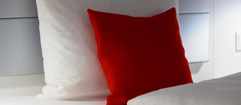 cama feita com uma almofada grande branca e uma almofada vermelha mais pequena a decorar