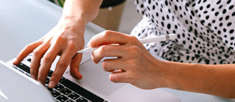 Mãos de mulher com blusa branca com bolas pretas com uma mão no computador e outra a segurar uma caneta como as pessoas canhotas.